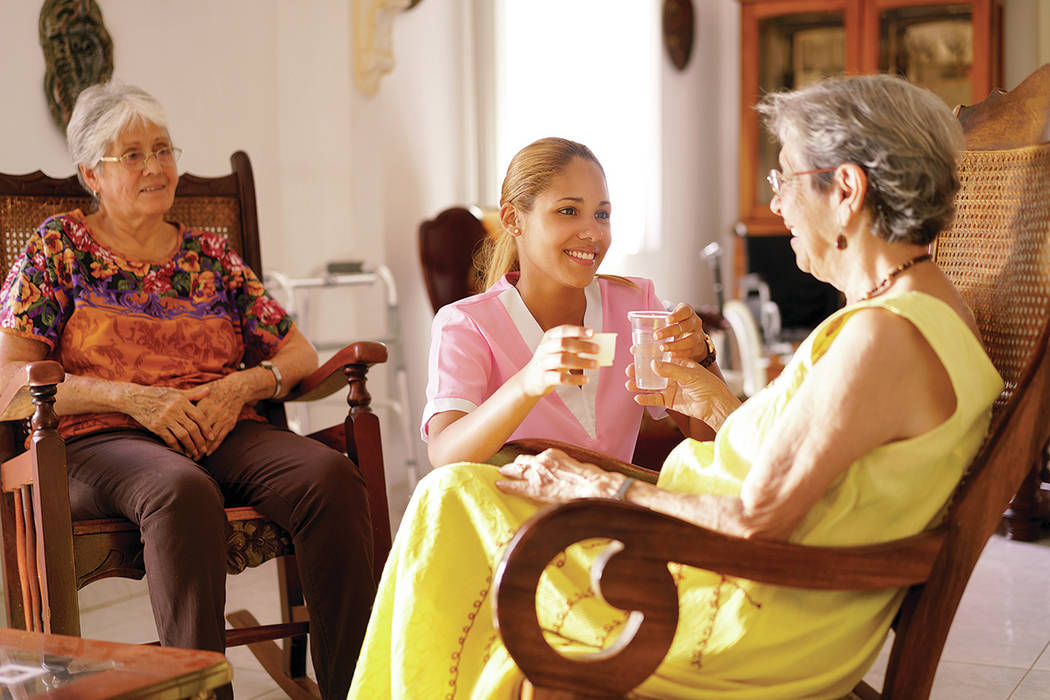 Personas mayores en un hospicio geriátrico: una joven hispana atractiva que como enfermera ayuda a una mujer mayor. Ella le da un vaso de agua y un medicamento recetado al paciente anciano. [ Fot ...